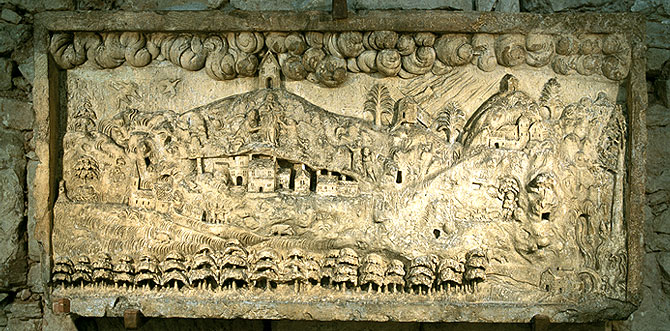 Steinernes Relief mit Darstellung einer hügeligen Landschaft, eines Dorfes mit Kirche, Bäumen und Feldern, an einer Steinmauer